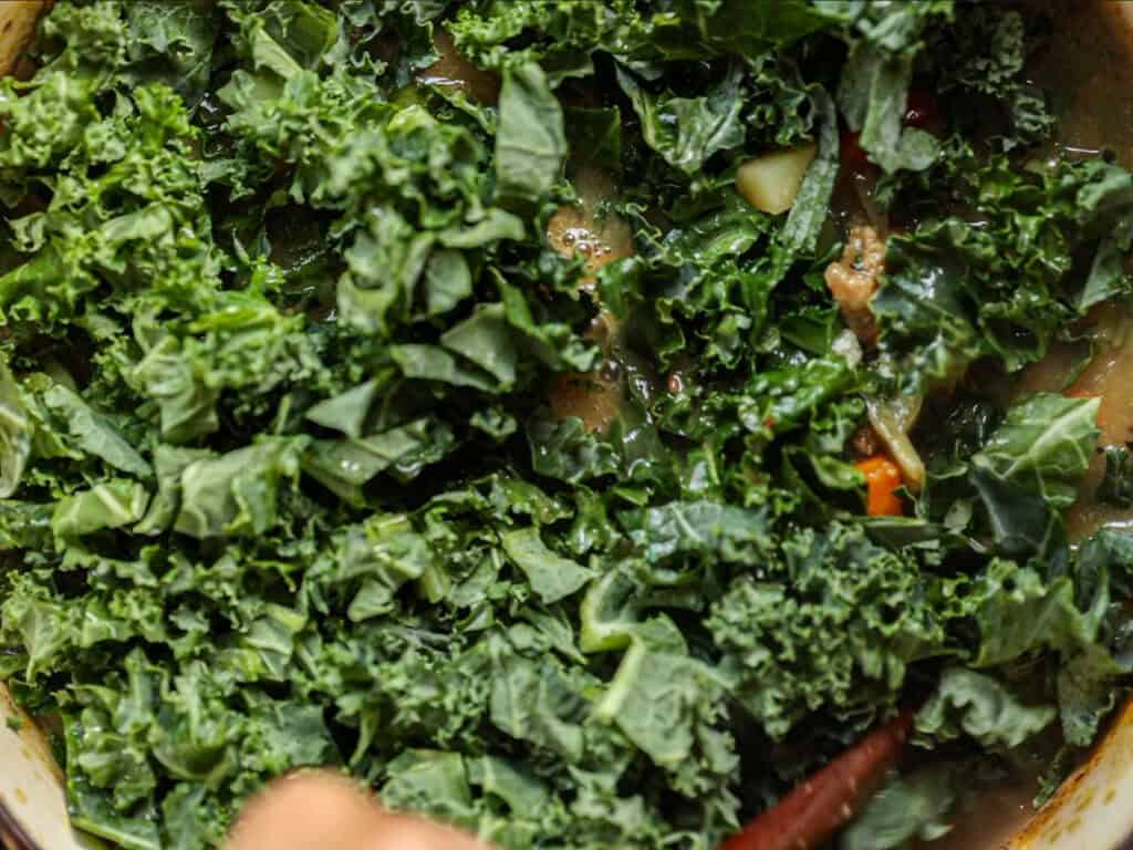Chopped fresh kale being cooked in a pot with a mixture of vegetables and broth visible underneath.